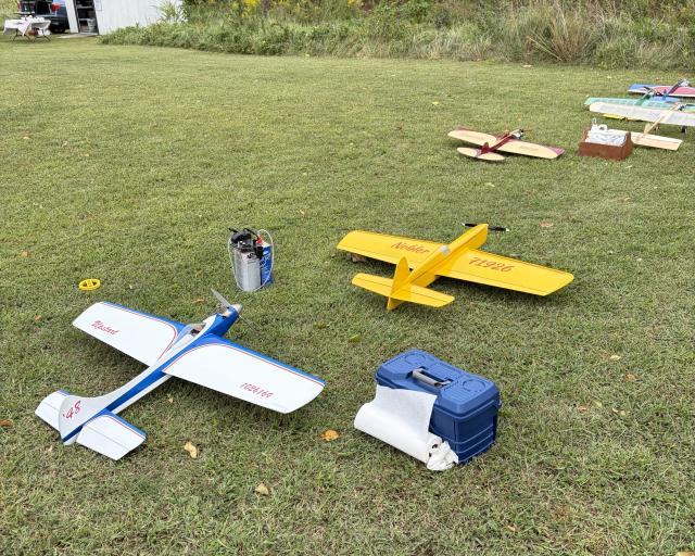 Several model airplanes and flight equipment are arranged on a grassy field before a flying demonstration or hobby event.