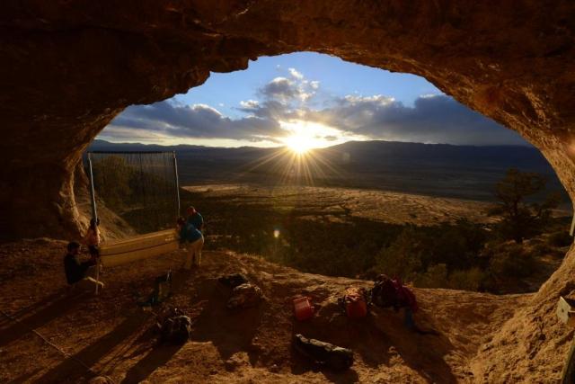 View from inside a large cave opening looking out over a broad desert valley at sunset. Sunlight streams through scattered clouds and over distant mountain ridges, creating a starburst effect on the horizon. Near the cave entrance, several people stand beside a tall mesh gate or barrier, with backpacks and equipment placed on the rocky ground. The cave’s rough stone walls frame the landscape beyond, highlighting the contrast between the shaded interior and the brightly lit valley outside.