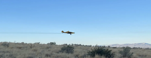 A small plane flies over a grassy, mountainous landscape. A trail of herbicide follows behind the plane, after being unloaded from the bottom.