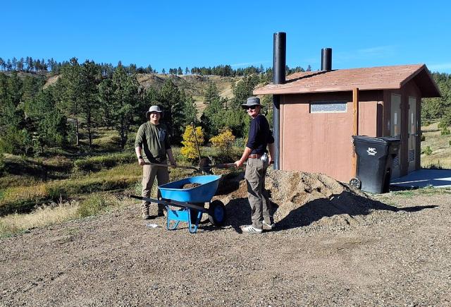 Two volunteers load a wheelbarrow from a pile of gravel