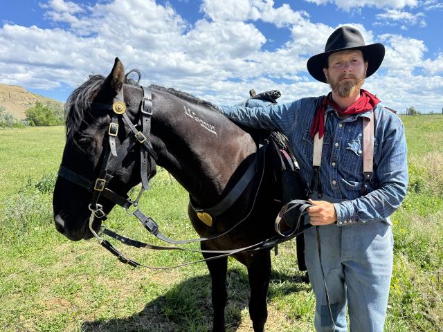 An individual in historical gear stands next to a black horse in a grassy field