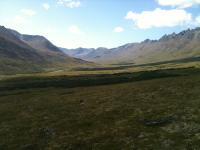 A valley full of green tundra stretches across the expanse between mountain ridges. The sky is blue and dots of clouds cast shadows on the valley.