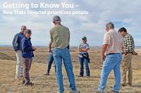 A group of seven people, standing, facing each other, conversing; short-grass prairie landscape with golden, late-season grasses; expansive, low-elevation hills in the background; partly cloudy sky.  