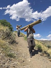 MCC crew members carrying timber over their shoulders down the trail on the side of a grassy hill. 