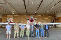 Eight members of the group stand in front of the plane. 