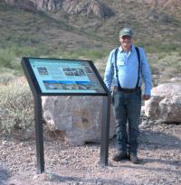 Person wearing suspenders and a ball cap standing next to an interpretive panel