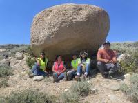 Five interns sitting under a large rock on a grassy surface. 