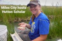 A young girl sits on the side of a creek, smiling at the camera, and holding a small fish in her hand. Text says Miles City hosts Hutton Scholar.