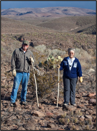Two hikers with stand in a rocky desert landscape with cacti and mountains in the background