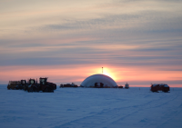 Snow-covered ground, with an orange sunset behind a large inflatable dome. Heavy equipment nearby.