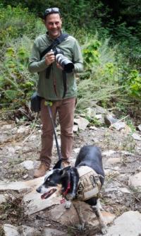 A man holding a camera stands next to a service dog.