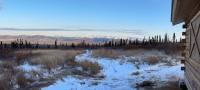 Picture of the view of the valley with the sun shining on the White Mountains' highlands in the distance. A few inches of snow covers the ground. The edge of the Moose Mountain Cabin frames the right side of the picuture.