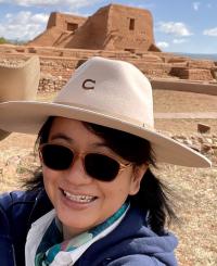 Hebin Lin, who is wearing a brimmed hat and sunglasses, smiles in front of Pecos National Park in New Mexico.