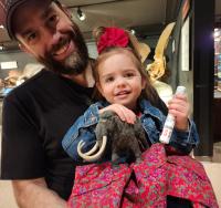 Man in black shirt holds young girl who holds a small stuff wooly mammoth