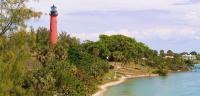 A photo of the Jupiter Ilet Lighthouse ONA. The tall orange lighthouse is seedn rising above the treetops to the left, while the bay, beach and more foliage are seen in the middle and right. 