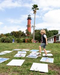 This photo shows a child walking among photos of lighthouses on the grass with a lighthouse and a palm tree in the background. 