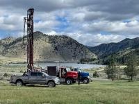a photo of trucks with well-digging equipment sit in a grassy field with a lake and mountain in the background under a mostly cloudy sky.
