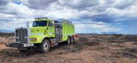 A light green wildland fire truck, or water tender, sits in the foreground under think grey clouds in a desert landscape that has been scorched black by a wildland fire.
