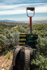 photo: small blackboard with writing propped on a backpack leaning on a shovel overlooking shrub-covered hills