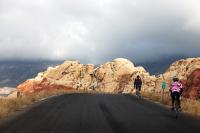 With thick clouds in the distance, two cyclists make their way up a smooth paved road bordered on all sides by cream colored boulders
