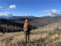a photo of a child wearing hunter orange and camouflage standing on a hill with sprawling mountain views under a blue sky with puffy white clouds.