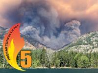 Thick smoke rises from a forested mountain landscape. An overlayed graphic displays the number 5 and a flame. 