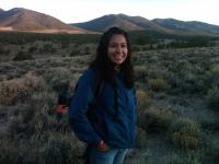 Vanessa Lacayo, smiling and wearing a blue jacket, stands in a field of sagebrush. Behind her is a line of small mountains, brownish green in color.