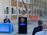 A woman in a tan suit stands behind a black podium in front of a brown apartment building that is under construction. A man in a suit sits at a table off to one side of the podium. The backs of the heads and shoulders of two other men seated in the audience are visible in the foreground.