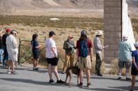 A uniformed person stands near an old building with people and a dog wander about.