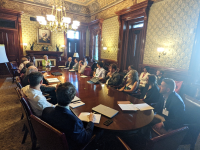Group of people sit around a table looking at Dr. Jane Lubchenco, located in a large room.