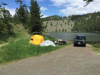 Alt Text: A photo showing a campsite next to Chopaka Lake. A black SUV is backed up to the lake on a dirt road, parked next to a picnic table and two tents.