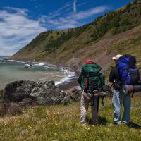 two backpackers view a mountain range rising above the ocean shore