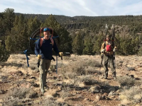 Two caucasian men pose outdoors in hiking clothes carrying metal stakes and wheels of wire on their backs.