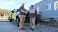 A Native American man poses shaking hands with a Caucasian man next to a green yellow truck