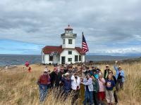 A group of volunteers are raising the American flag. They are standing in tall yellow grass with a white lighthouse in the background. 