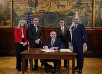 Interior Secretary Doug Burgum sits at a table and smiles after signing documents. Utah Senator John Curtis is to his right, Acting BLM Director Jon Raby and Utah Governor Spencer Cox are to Burgum's right.
