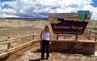 A woman in sunglasses stands facing the camera and smiling next to a sign with the BLM logo. The sign reads “Gunnison Gorge National Conservation Area.” Behind her, beyond an area of shrubland, are light brown and green mountains beneath a sky with large white clouds and patches of blue. 