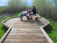 A photo showing a wooden boardwalk leading away from the viewer. A teenage boy sits on a wooden bench on the widened part of the boardwalk with his younger sister who is sitting in a mobility device.