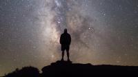 A photo showing the silhouette of a man standing on a high point of ground looking up at the night sky, which is a blend of gray and beige, dotted heavily with stars. The Milky Way is positioned vertically in the center of the photo.  