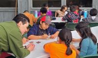Photo. Uniformed Ranger leans over a table assisting students with a paint making activity. Students hold brushes and are painting on tan colored rocks. Ten students can be seen in the image, sitting around tables in the daytime. 