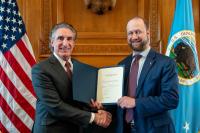 Two men in suits shake hands while holding a certificate in a formal wood-paneled room with U.S. flags behind them.