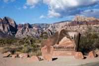 A sandstone sign reading "Red Rock Canyon National Conservation Area - Bureau of Land Management" stands in front of red and orange-striped rock formations and yucca plants under a blue sky with white clouds.
