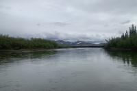 A river sits beneath an overcast sky, its smooth surface interrupted by little ripples. Green vegetation reflects on either side of the water, and rolling hills with patches of snow are visible in the distance.