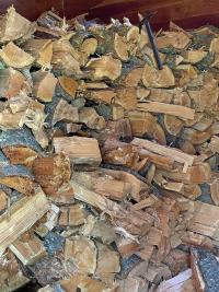 Photo. Cut firewood; a pile of randomly positioned firewood in the foreground; stacked firewood in the background; a black-colored hatchet hangs from the top of one piece of wood in the stack.