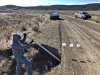 Photo. Daytime, sunny, partly cloudy, blue skies. A gravel road extends from the foreground into the distance through a dry prairie landscape. A left, a man holds a long, white measuring stick to determine the depth of a hole in the road. 