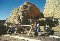 A group of volunteers works together to build a wooden fence structure in a desert landscape with tall sandstone cliffs rising in the background.