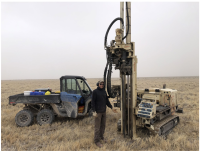 A person stands next to a tall, slender machine several times his own height. An ATV and small, tracked machine sit in the background.