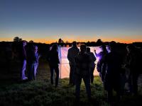 A group of people gathers around a brightly lit screen in a field at dusk, with a colorful sunset in the background.