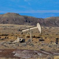An oil pumpjack operates in a dry desert landscape with sparse vegetation, rocky outcrops, and distant mountains in the background.
