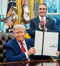 President Trump sits at his desk and holds up one of the energy executive orders he signed with Secretary of the Interior Burgum standing behind him clapping in the oval office.